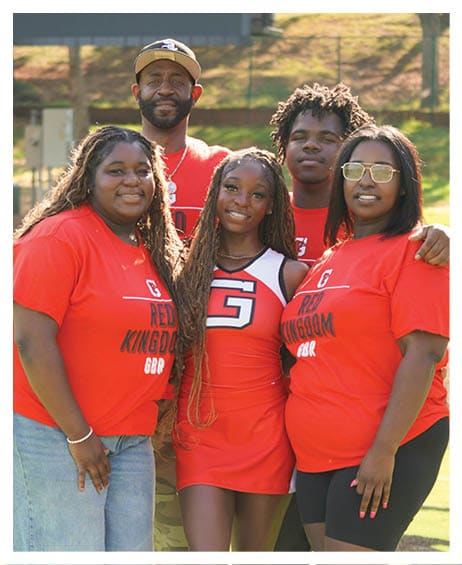 Five people wearing red shirts and one person in a cheerleading uniform are standing closely together outdoors with a scoreboard and trees in the background. Description generated by AI