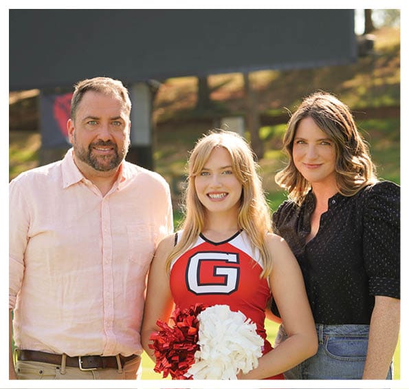 Three adults stand outdoors with one wearing a red cheerleading outfit holding red and white pom-poms while the others wear casual clothing and smile. Description generated by AI
