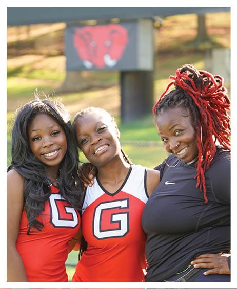 Three people smiling and posing together outdoors with two wearing matching red sports uniforms and one in a black athletic top with red dreadlocks. Description generated by AI