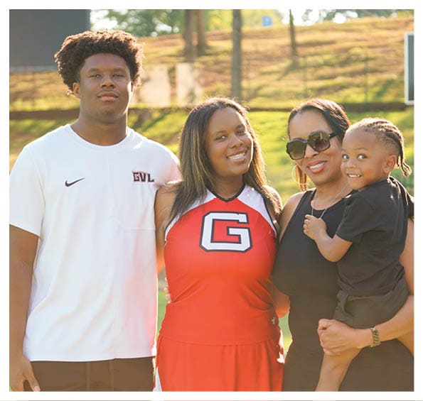 Four people stand outdoors in sunlight with one person holding a child, all smiling and dressed casually with one wearing a sports uniform. Description generated by AI