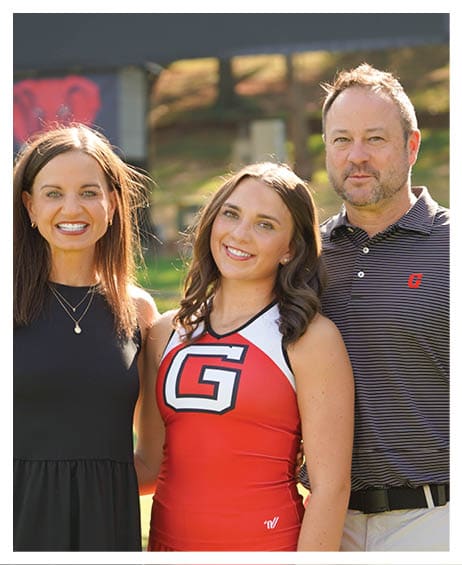 Three adults stand closely together outdoors with one wearing a red cheerleading uniform and the others dressed in casual clothing, all smiling in natural sunlight. Description generated by AI