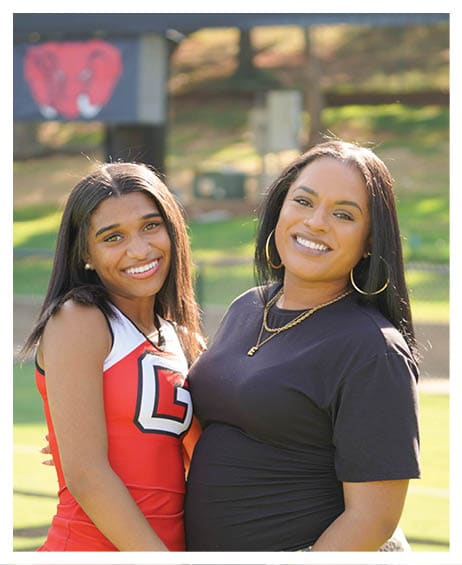 Two people with long hair smile warmly while standing close together outdoors on a sunny day, one wearing a red and white cheerleading outfit and the other wearing a black shirt with large hoop earrings and a necklace. Description generated by AI