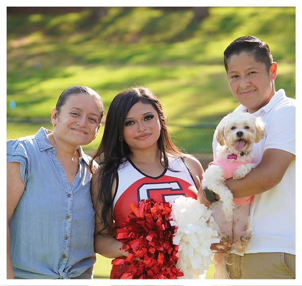 Three people stand outdoors on grass with one holding a small dog wearing a pink outfit while another person wears a cheerleading uniform and holds red and white pom-poms. Description generated by AI