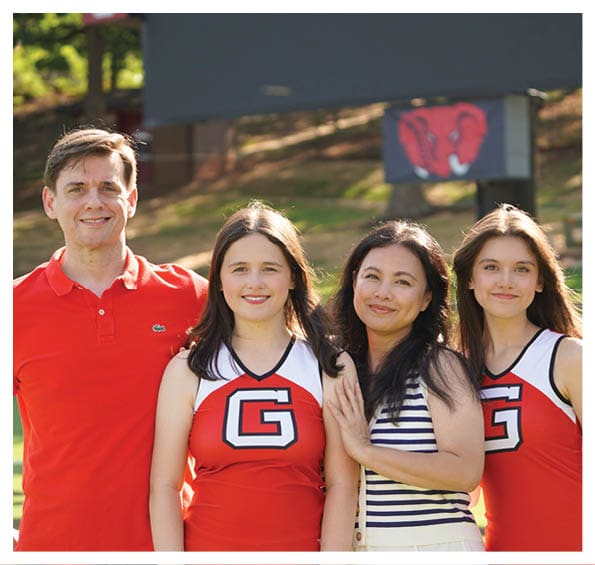 Four people standing closely together outdoors with two wearing red cheerleading uniforms and one wearing a red polo shirt, all smiling. Description generated by AI