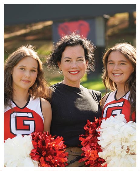 A person in a red and white cheerleading uniform holding red and white pom-poms stands next to another person wearing a black shirt outdoors on a sunny day. Description generated by AI