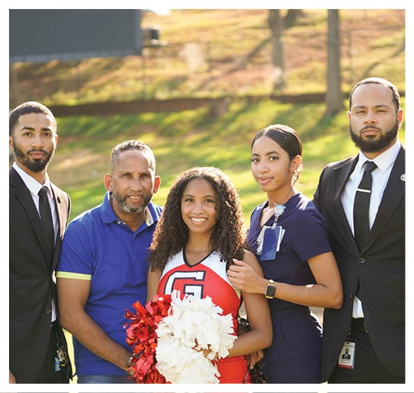 Four people stand closely together outdoors with one person in a cheerleading outfit holding red and white pom-poms while the others wear casual and formal clothing. Description generated by AI
