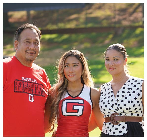 Two people are smiling outdoors with one wearing a black dress and the other wearing a red cheerleading uniform with a large letter G on the front. Description generated by AI