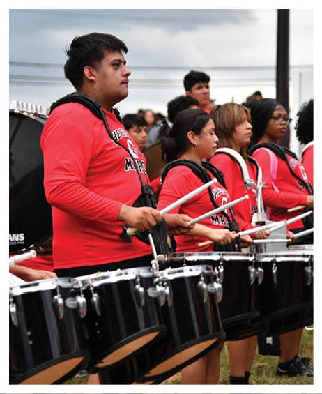 A line of people wearing matching red shirts are playing black marching drums outdoors on grass. Description generated by AI