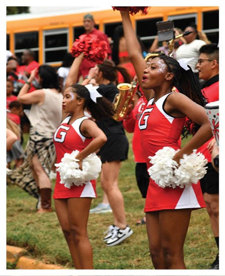 Two adults in red and white cheerleading uniforms with pom-poms perform outdoors while a band plays in the background. Description generated by AI