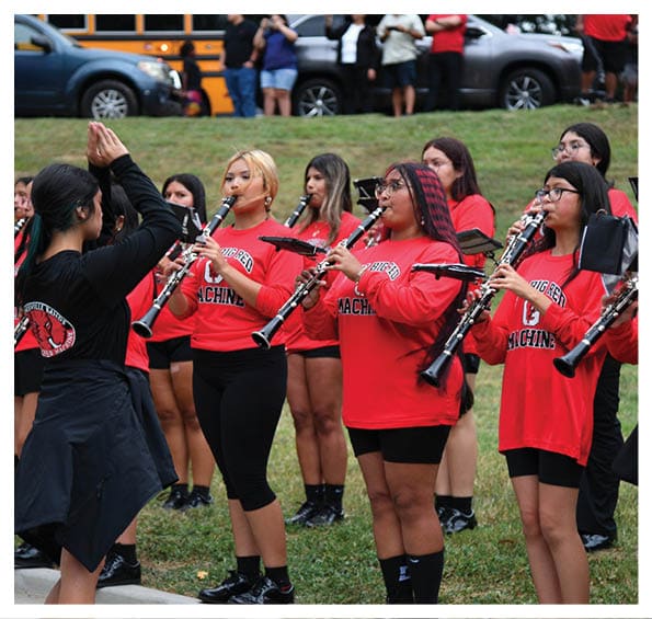 Adults wearing red shirts with “Big Red Machine" printed on them are playing clarinets outdoors while a person in black conducts them. Description generated by AI