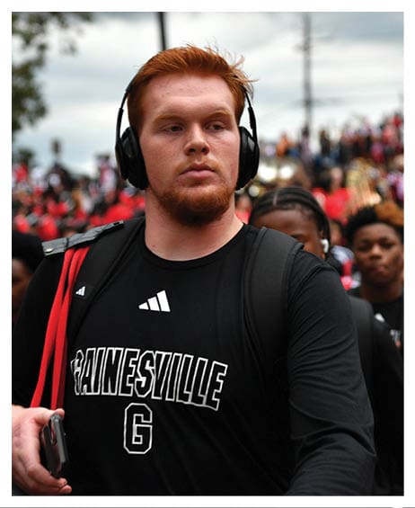 A person with short red hair wearing headphones and a black Gainesville shirt is walking outdoors with a crowd in the background. Description generated by AI