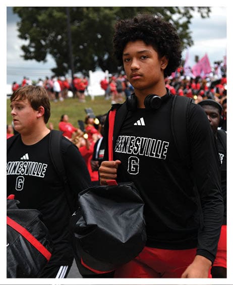 Two people wearing black Gainesville shirts and carrying bags walk outdoors with a crowd and trees in the background. Description generated by AI