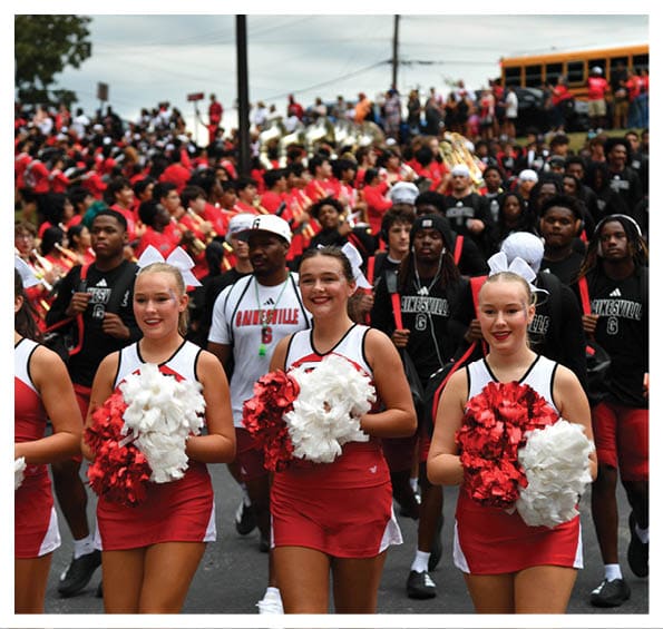 A group of people in red and white cheerleading uniforms holding pom-poms leads a marching band dressed in black and red down a street during a parade or event. Description generated by AI