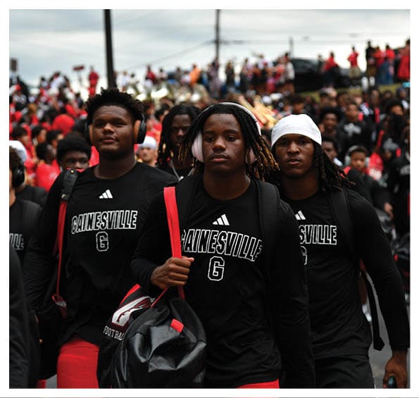 Three people wearing matching black Gainesville shirts and carrying bags walk forward with a crowd of people in the background. Description generated by AI