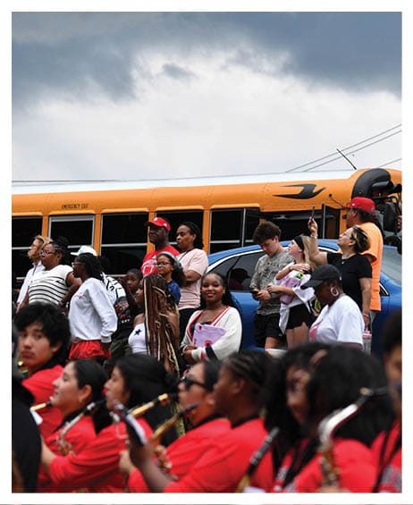 A group of people stand and watch while a band dressed in red uniforms plays instruments in front of a yellow school bus under a cloudy sky. Description generated by AI