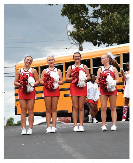 Four people in cheerleading uniforms holding red and white pom-poms stand on a street with a yellow school bus and trees in the background. Description generated by AI