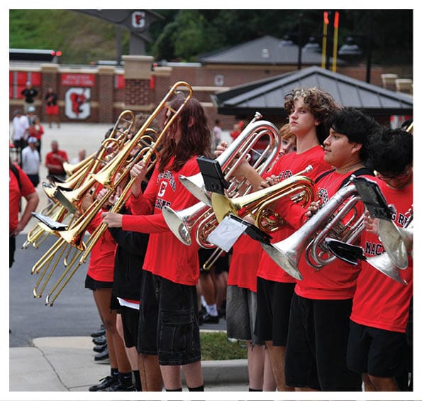 A group of people wearing red shirts are standing outdoors playing brass instruments in a band formation near a sports stadium. Description generated by AI