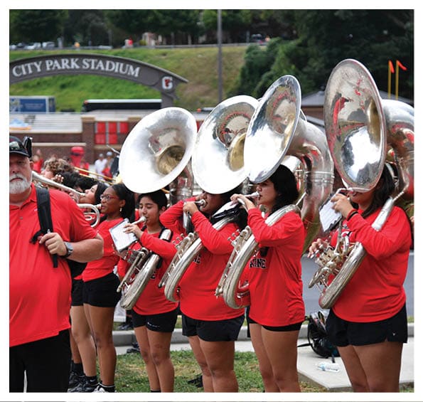 A group of people wearing matching red shirts and black shorts are playing large brass instruments outdoors near a stadium sign that reads “City Park Stadium." Description generated by AI
