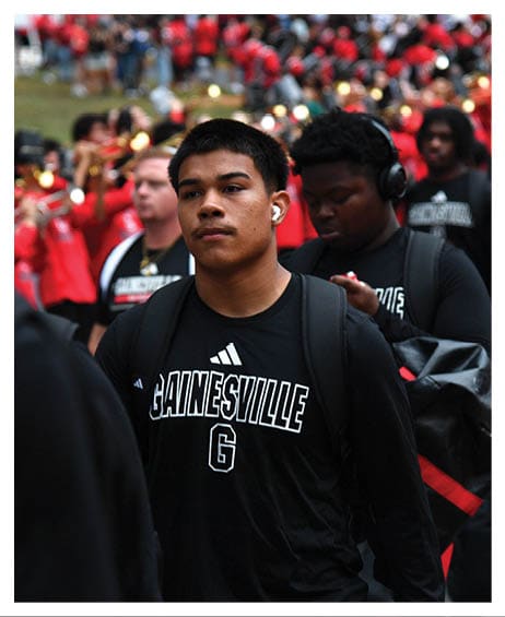 A person wearing a black Gainesville shirt and backpack walks among a crowd with some people playing brass instruments in the background. Description generated by AI