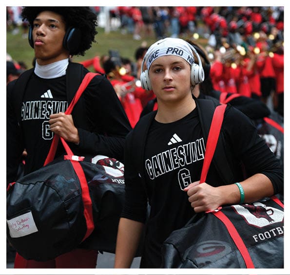 Two people wearing headphones and black Gainesville sports shirts carry large black and red duffel bags with a marching band visible in the background. Description generated by AI