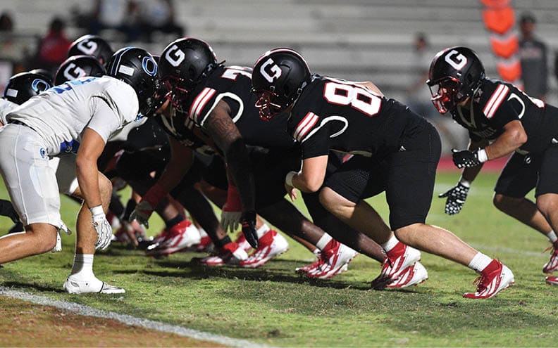 A group of people in black football uniforms with helmets are lined up on a field, preparing for a play. Description generated by AI