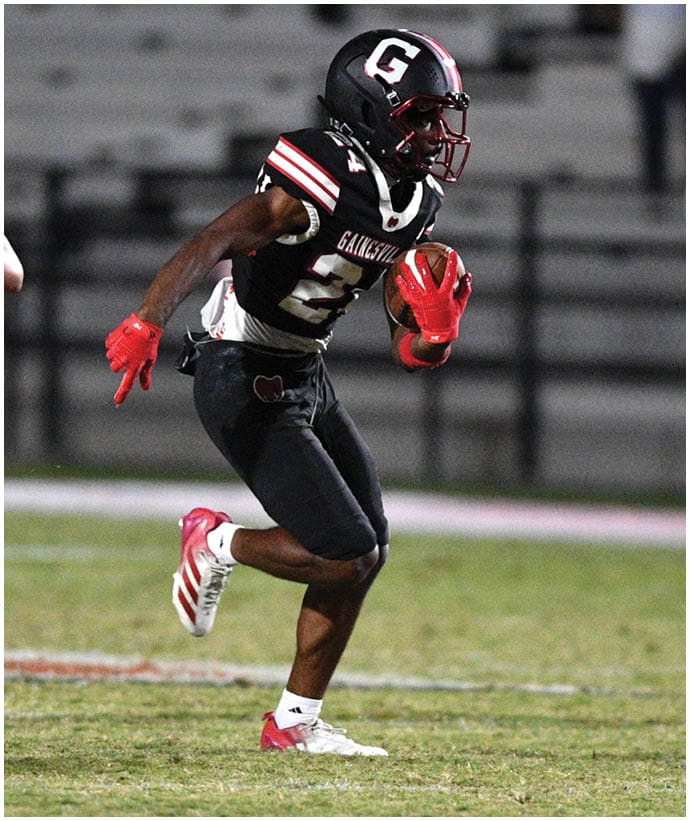 A person in a black football uniform with red gloves is running with the ball while being chased by a person in a white football uniform on a grassy field with empty bleachers in the background. Description generated by AI