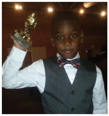 A child dressed in a white shirt, gray vest, and colorful bow tie is holding up a small trophy in a dimly lit indoor setting. Description generated by AI