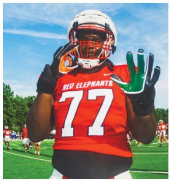 A person wearing a red football jersey with the number 77 and the words “RED ELEPHANTS" is standing on a football field making hand gestures while wearing a white helmet and gloves. Description generated by AI