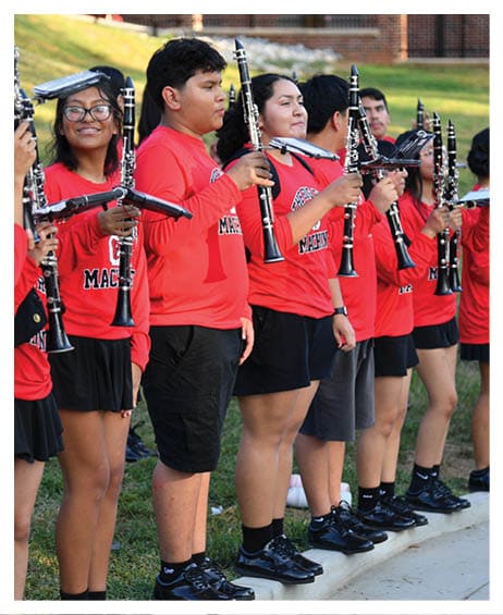 A group of people wearing matching red shirts and black shorts stand in a line outdoors holding clarinets. Description generated by AI