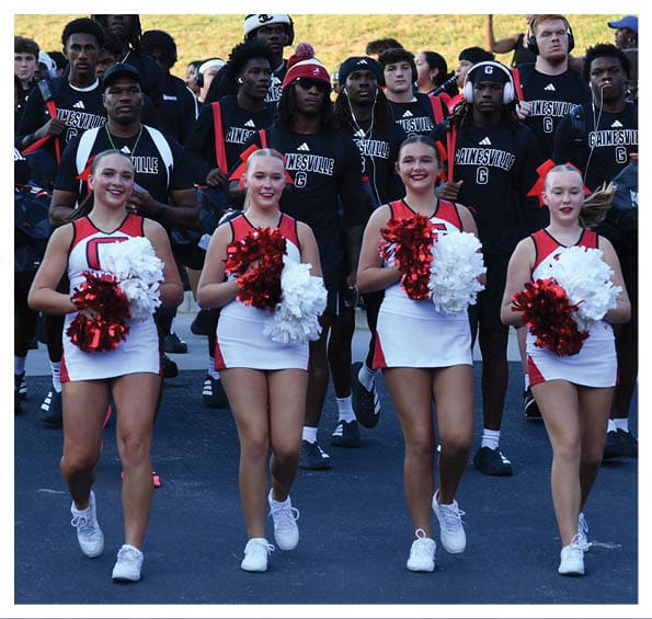 A group of adults in matching athletic uniforms is walking together on a paved road with four adults in cheerleading outfits holding pom-poms leading the group. Description generated by AI