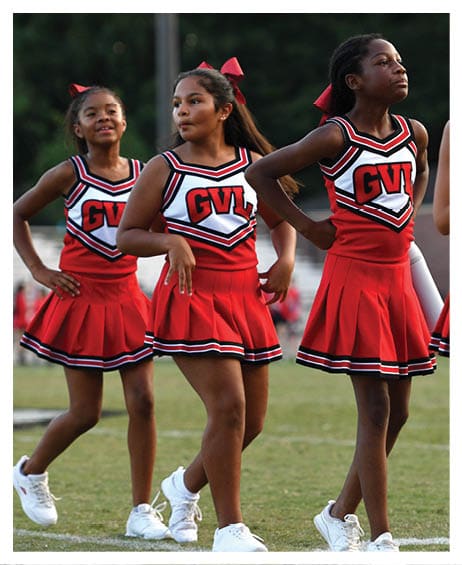 Three children in red cheerleading uniforms with “GVL" on the front stand on a grassy field, appearing focused and ready to perform. Description generated by AI