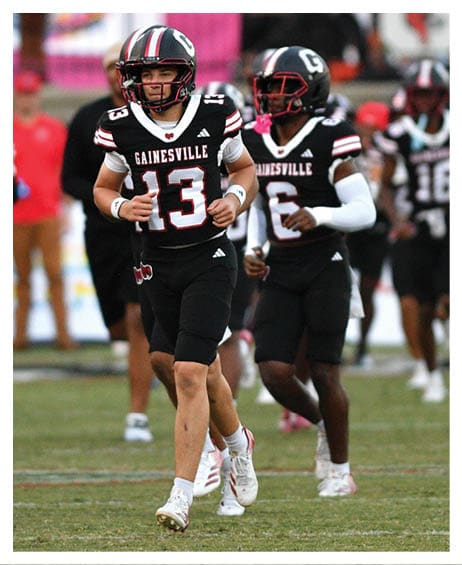 A group of football players in black uniforms with “Gainesville" on their jerseys are running on a grassy field with a brick building and a pink tent in the background. Description generated by AI