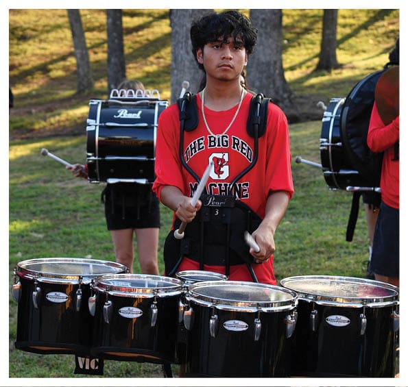 People wearing red shirts are playing various marching band instruments outdoors on a grassy area with trees in the background. Description generated by AI