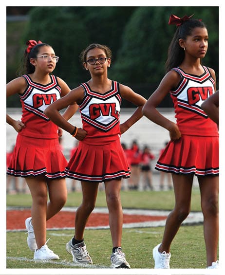 Three children wearing red and white cheerleading uniforms with “GVL" on the front stand on a grassy field with their hands on their hips. Description generated by AI
