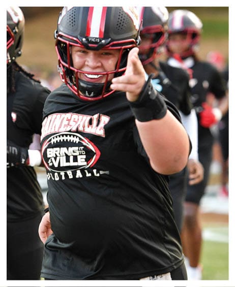 A person wearing a football helmet and black sportswear with “BRING IT VILLE FOOTBALL" printed on the shirt is smiling and pointing towards the camera on a football field with other players in the background. Description generated by AI