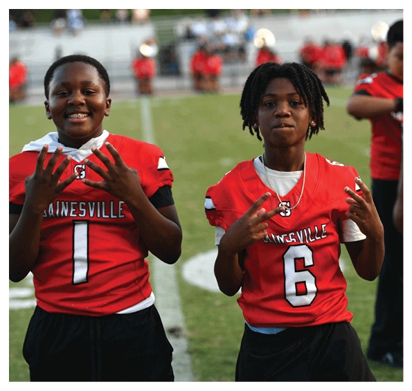 A person wearing a red sports jersey with the number six is standing on a grassy field making hand gestures while other people in similar jerseys stand in the background. Description generated by AI
