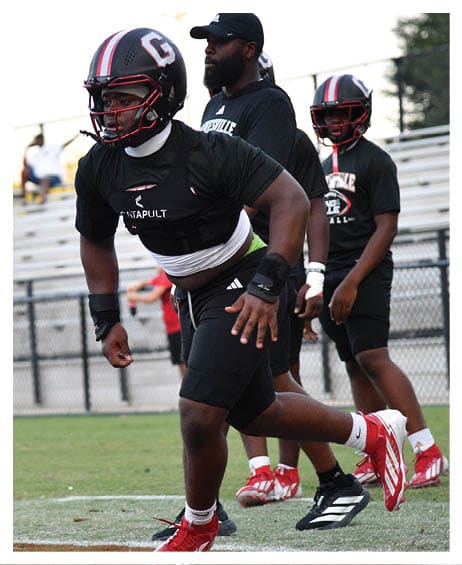 A person wearing a football helmet and athletic gear is running on a field while two other people stand nearby, one wearing a cap and the other also in football gear. Description generated by AI