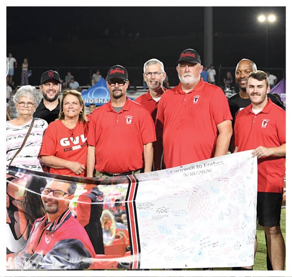 A group of people wearing red shirts and casual clothing stand together on a sports field holding a large banner with a photo and handwritten messages, under stadium lights at night. Description generated by AI
