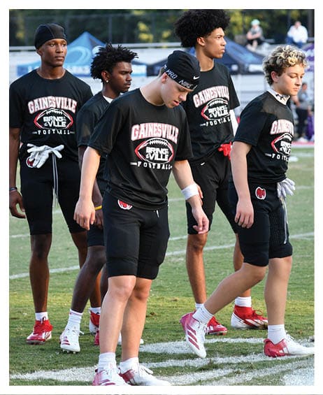 Five people wearing matching Gainesville football shirts and athletic shorts stand on a grassy field, appearing focused and preparing for an activity. Description generated by AI