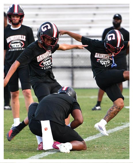 Two people in black athletic clothing are on a grassy field, with one kneeling and holding a football while the other is in motion, possibly kicking. Description generated by AI