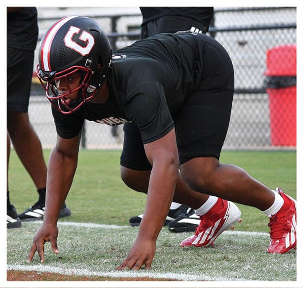 A person wearing a black helmet and black athletic clothing is crouched in a football stance on a grassy field with others standing nearby. Description generated by AI