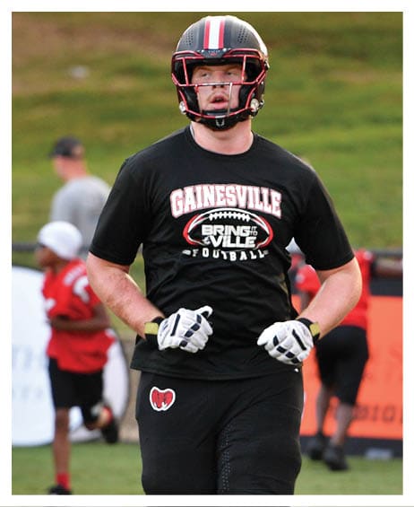 A person wearing a football helmet, gloves, and knee brace is running on a football field with a black Gainesville football t-shirt and black pants. Description generated by AI