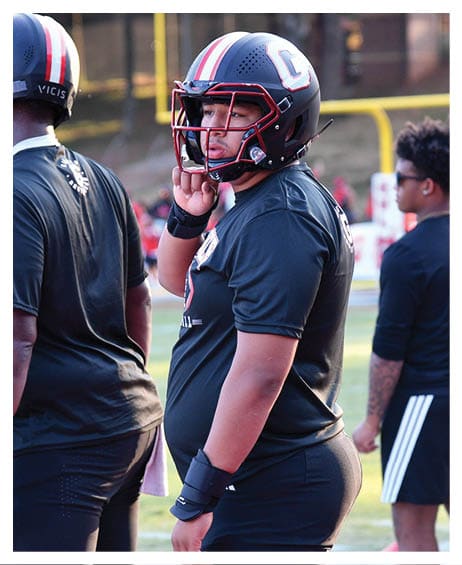 Two people wearing football helmets and black athletic clothing stand on a field while another person in black sportswear stands nearby. Description generated by AI