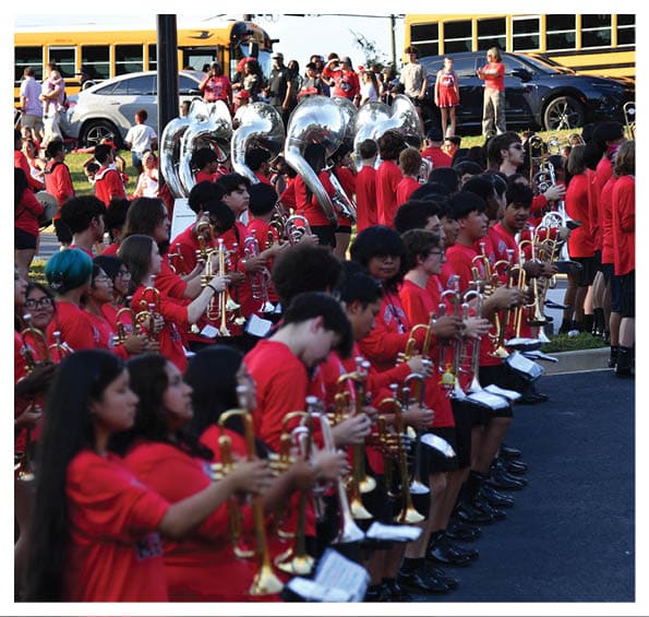 A large group of people wearing red shirts are standing outdoors playing brass instruments in a band formation with school buses and cars in the background. Description generated by AI