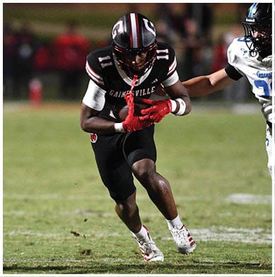 A person wearing a black sports uniform with red gloves is running on a grassy field while holding a football, with other players nearby. Description generated by AI