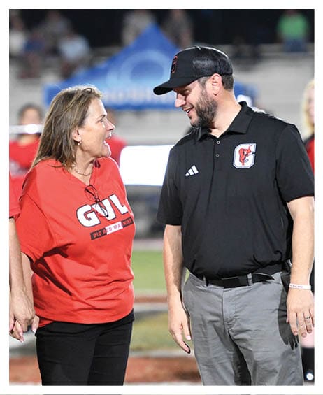 Three adults stand on a sports field holding hands and smiling, with one wearing a red shirt and the others wearing a red polo and a black polo. Description generated by AI