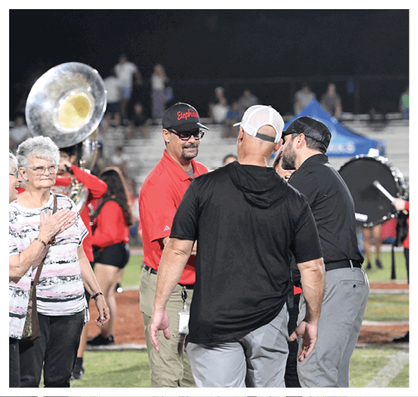 Adults wearing casual clothing are standing and interacting on a sports field with a marching band playing instruments in the background. Description generated by AI