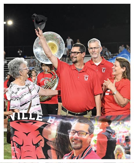 A group of people wearing red shirts stand together at an outdoor event with one person raising a hat and a banner featuring a smiling person and a red dragon logo in front of them. Description generated by AI