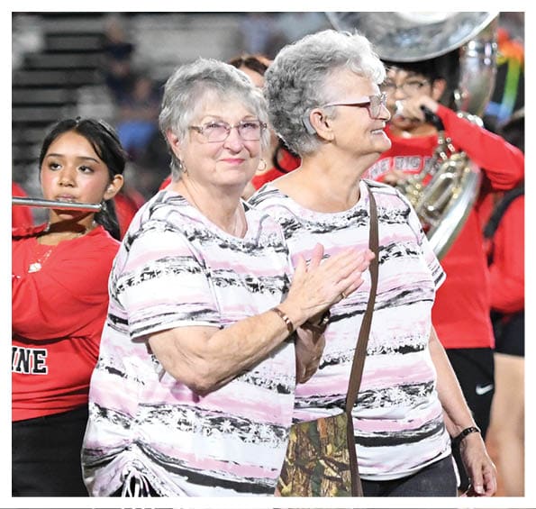 Two adults wearing red shirts stand on a sports field with a marching band playing instruments in the background. Description generated by AI