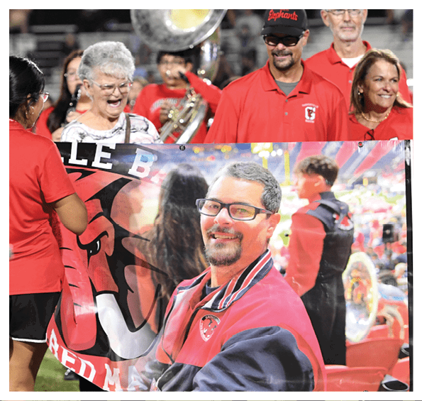 People wearing red shirts and hats are smiling and holding a large banner featuring a person in a red and black jacket with glasses, set in a lively outdoor event with musical instruments in the background. Description generated by AI